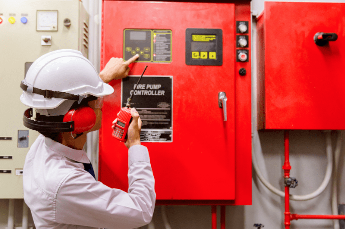 A man in white hard hat and safety glasses is pointing to the fire pump control panel.